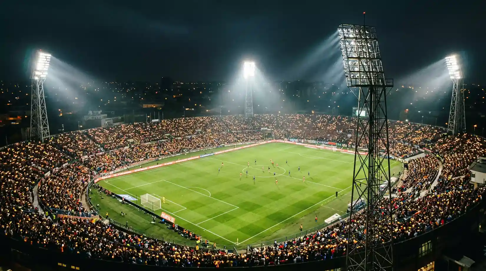 Fodboldstadion oplyst af projektører om natten med spillere på banen og mørk himmel